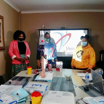 Three people standing in front of a screen with a logo, in a room with a table and various items.  VeinTrain flat-pack vein simulator for phlebotomy and cannulation training
